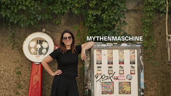 Lisa Gadenstätter in schwarzer Kleidung lehnt neben einem Automaten und einer historischen Waage vor einer begrünten Mauer, darüber steht „Mythenmaschine“. Hier bekommt man statt Kaugummis und Spielzeug Mythen. Doch welche spuckt der Automat aus? 