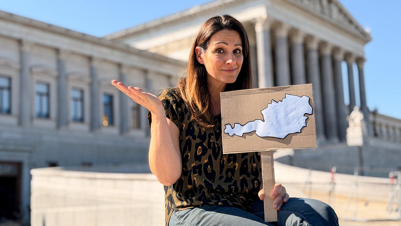 © ORF/BFILM Lisa Gadenstätter sitzt vor dem österreichischen Parlament und hält ein Schild mit der Umrisskarte Österreichs in die Kamera.