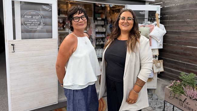 Ulla Kramar-Schmid und Tamara Hanl stehen lächelnd vor dem Geschäft „Glücksladen“, dessen Eingang mit Deko- und Geschenkartikeln gestaltet ist.