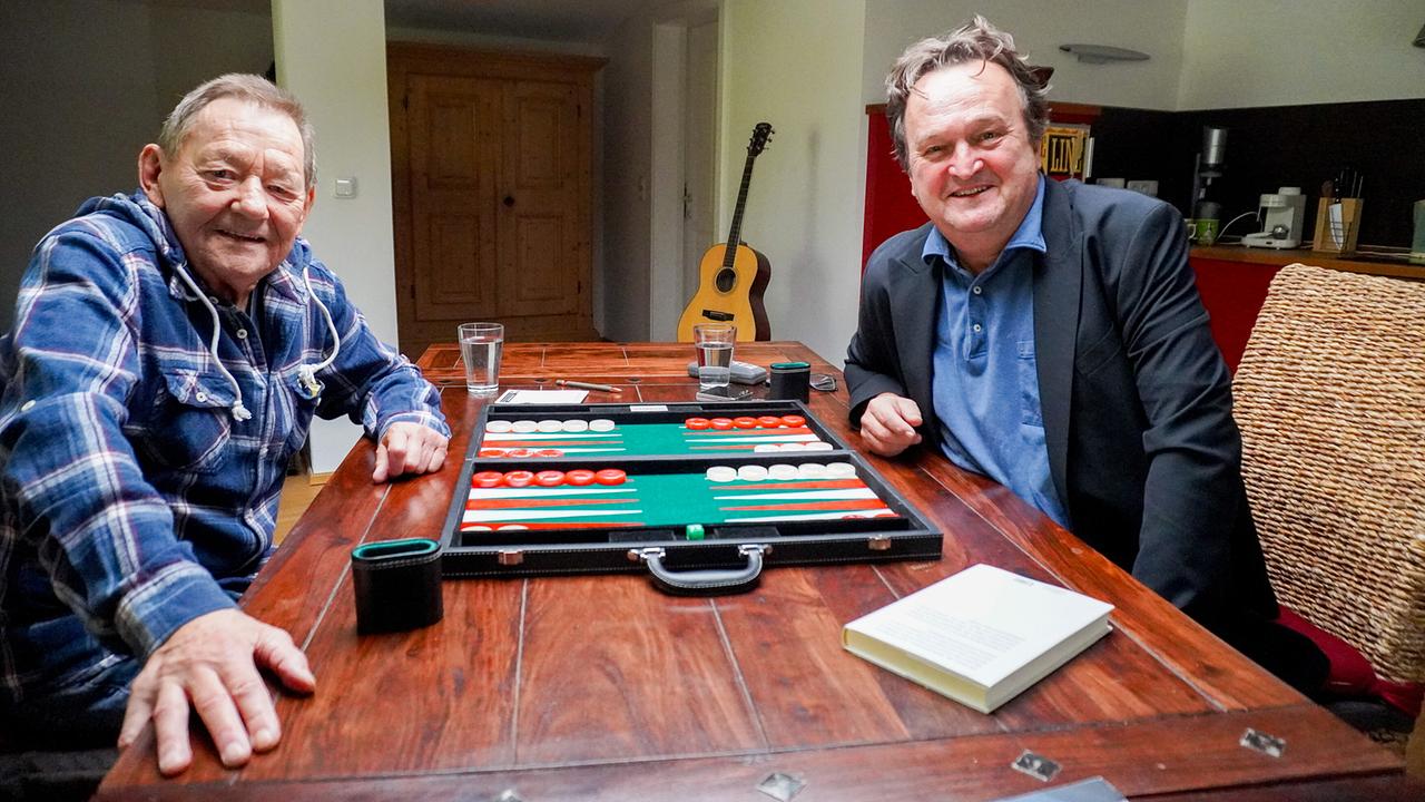 © ORF/Neulandfilm/Simon Schennach Wolfgang Ambros und Hanno Settele sitzen an einem rustikalen Holztisch mit einem Backgammon-Brett in Ambros Haus in Waidring in Tirol.