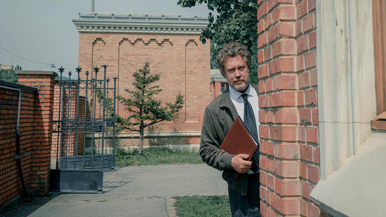 © ORF/Amazon/MGM Nicholas Ofczarek schaut als Heli Wondratschek hinter einer Ecke eines historischen Backsteingebäude hervor und hält eine Aktenmappe in der Hand