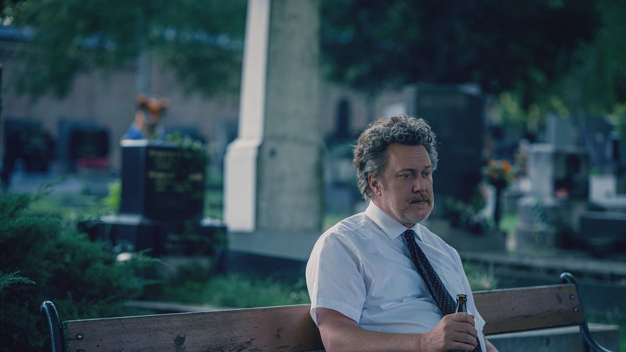Ein Mann sitzt auf einer Bank am Friedhof, hält eine Bierflasche in der Hand und blickt nachdenklich zu Boden