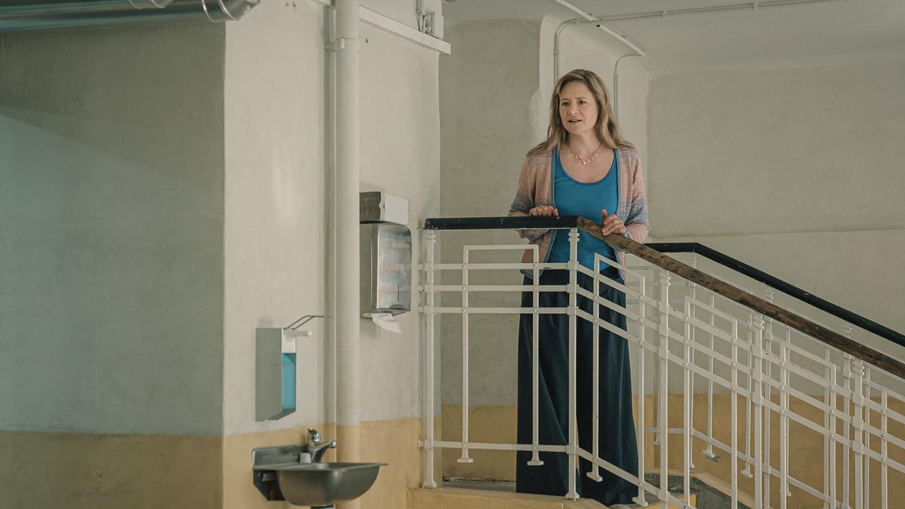 Eine Frau mit mittelblondem langen Haar, mit einem blauen Top, Strickjacke und dunklem Rock bekleidet, steht am Geländer eines historischen Stiegenhauses