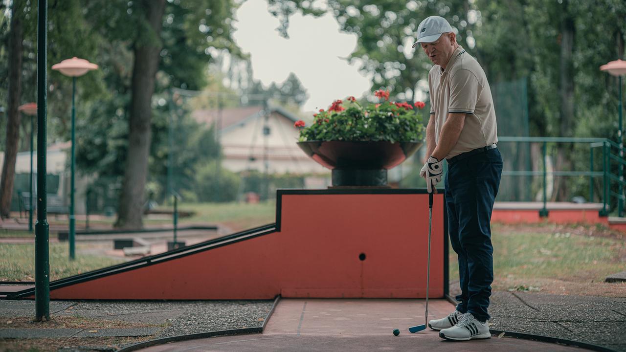 © ORF/Amazon/MGM Ein Mann mit beigen Polo-Shirt, dunkler Hose und Schirmmütze steht auf einer Minigolf-Bahn und zielt mit seinem Schläger auf den Ball