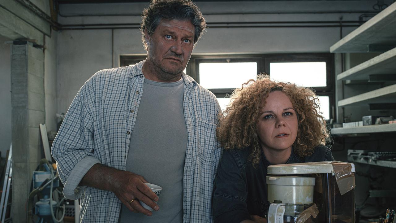 Ein Mann in T-Shirt und offenem karierten Hemd und eine Frau mit dunklen Oberteil stehen an einem Tisch mit Kaffeemaschine - beide blicken zweifelnd in dieselbe Richtung