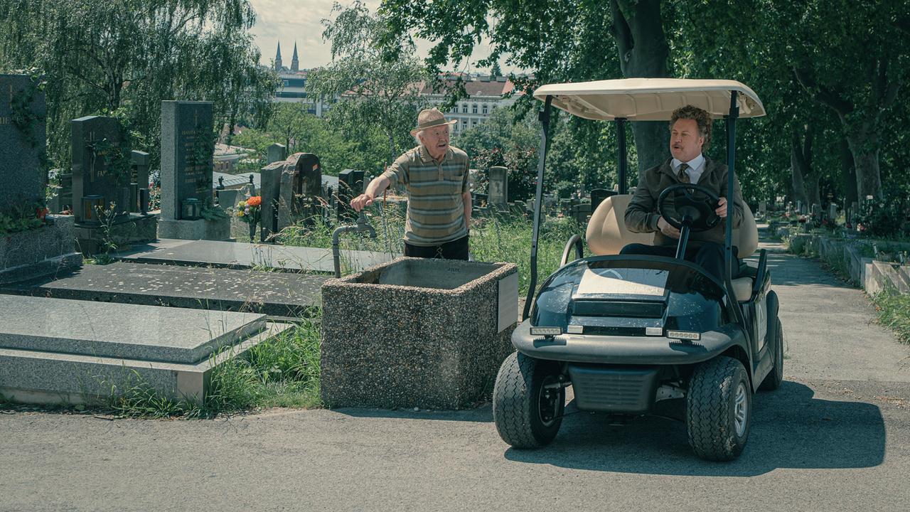 Ein Mann in grauer Jacke, Hemd mit Krawatte fährt in einem elektrischen Golfwagen über den Friedhof, ein älterer Mann steht am Wasserhahn und sieht zu ihm hin