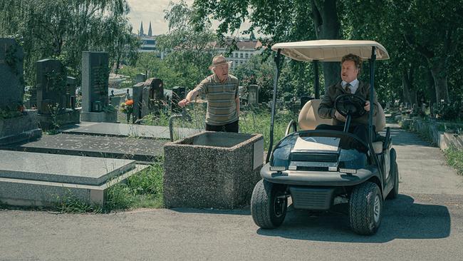 © ORF/Amazon/MGM Ein Mann in grauer Jacke, Hemd mit Krawatte fährt in einem elektrischen Golfwagen über den Friedhof, ein älterer Mann steht am Wasserhahn und sieht zu ihm hin