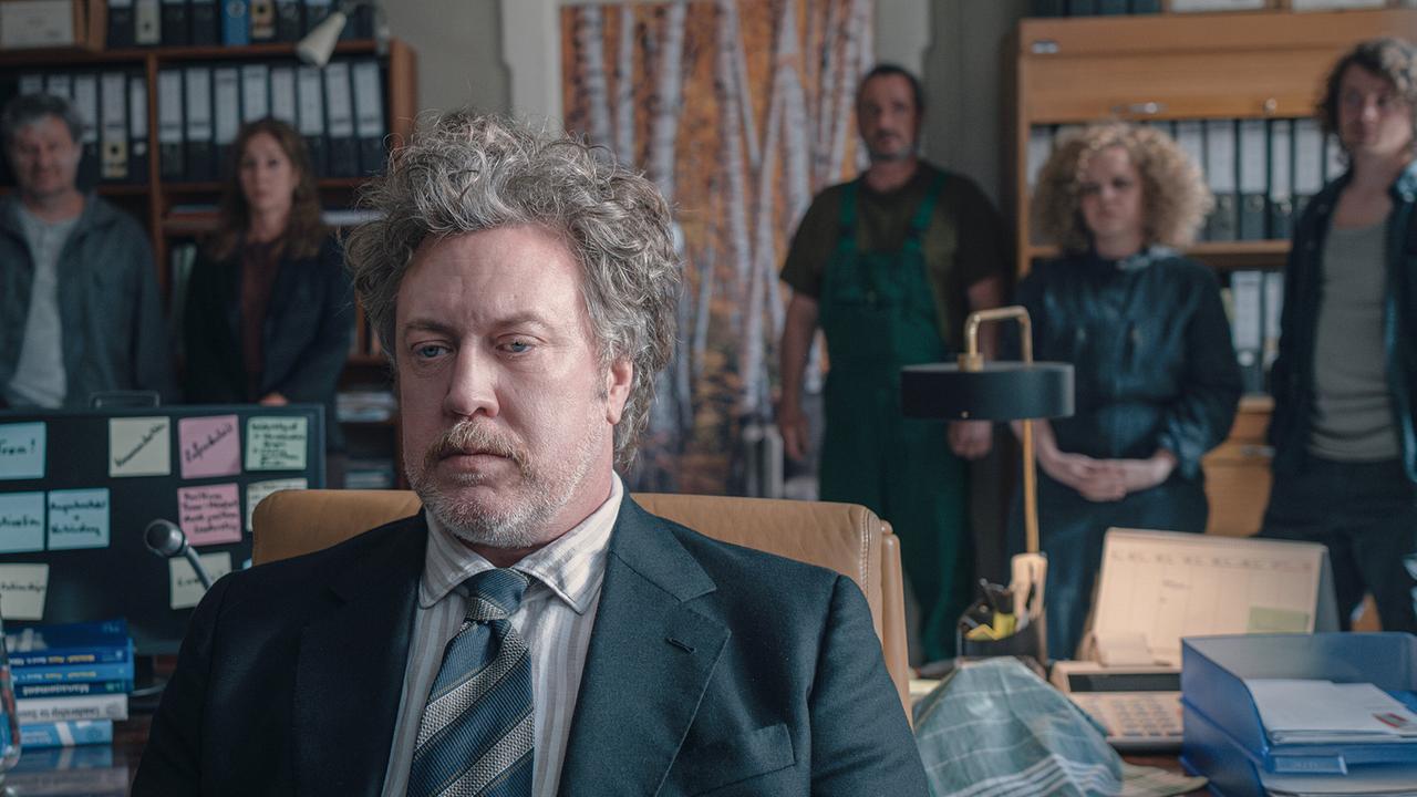 © ORF/Amazon/MGM Ein Mann mit grauem, wirren Haar und Bart sitzt im Anzug, gestreiftem Hemd und Krawatte im Büro - hinter ihm stehen fünf Personen vor vollen Aktenschränken