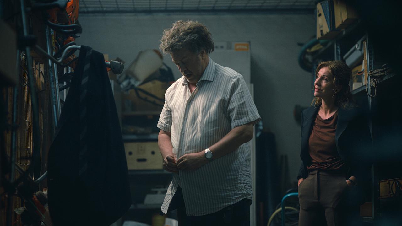 © ORF/Amazon/MGM In einem Lagerraum knüpft sich ein Mann mit wirrem grauen Haar das Hemd zu, eine dunkel bekleidete Frau steht hinter ihm und spricht mit ihm