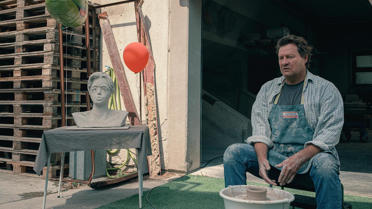 © ORF/Amazon/MGM Ein Mann mit kariertem Hemd und Schürze sitzt an einer Töpferscheibe, auf einem Tisch daneben eine Büste einer Frau und zwei bunten Luftballons