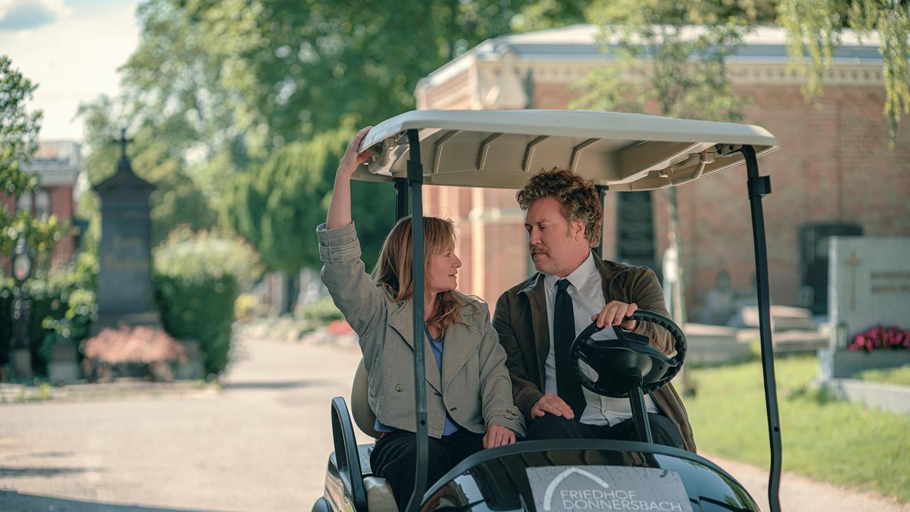 Eine Frau und ein Mann fahren gemeinsam in einem elektrischen Golfwagen über den sonnigen Friedhof