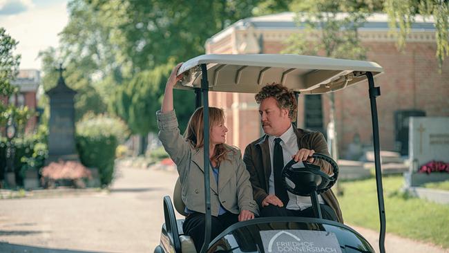 © ORF/Amazon/MGM Eine Frau und ein Mann fahren gemeinsam in einem elektrischen Golfwagen über den sonnigen Friedhof