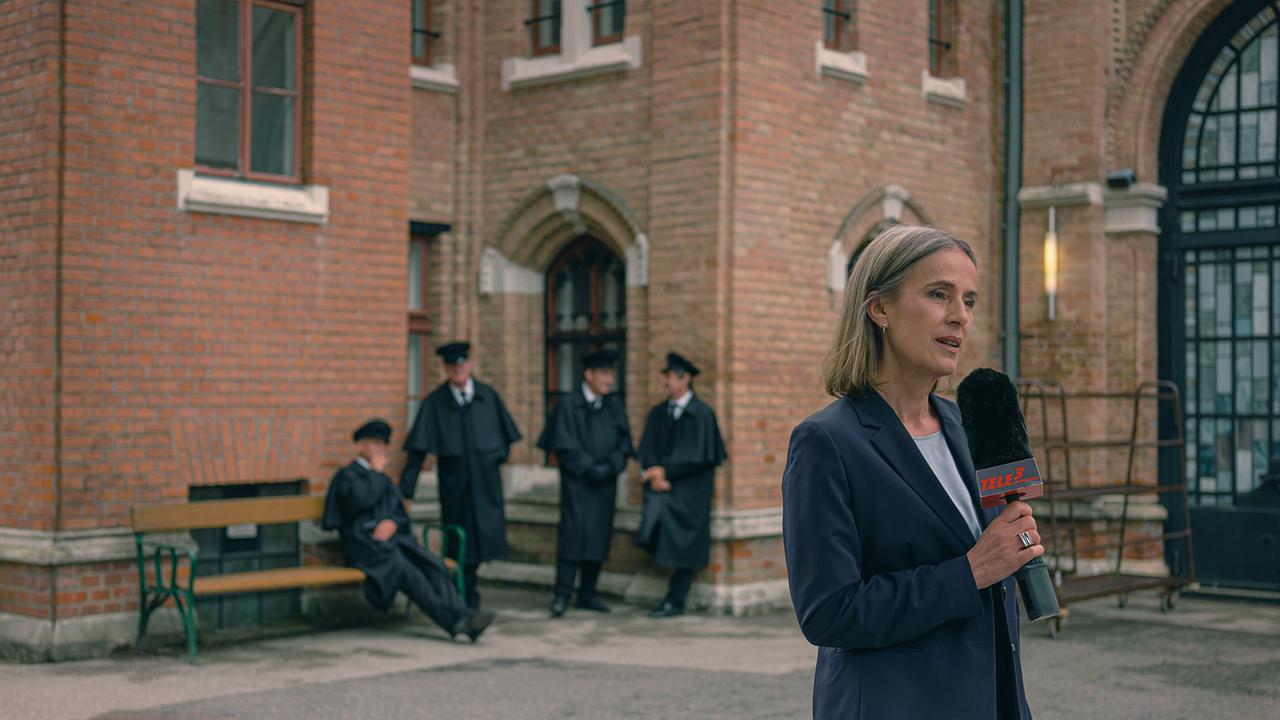 Eine Frau mit Mikrofon steht vor einem historischen Backsteinhaus, im Hintergrund sind vier Männer in schwarzer Kleidung von Friedhofsbeamten zu sehen