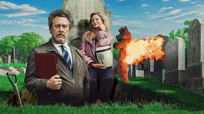 © ORF/Amazon/MGM Nicholas Ofczarek im Anzug steht in einem Grab mit einer Urne in der Hand, hinter ihm lehnt Julia Jentsch sitzend an einem Grabstein, dahinter läuft eine Person in brennendem Tierkostüm über den Friedhof.