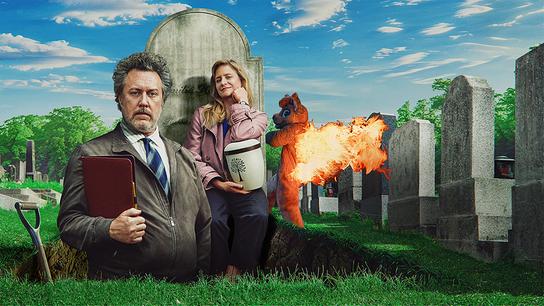 Nicholas Ofczarek im Anzug steht in einem Grab mit einer Urne in der Hand, hinter ihm lehnt Julia Jentsch sitzend an einem Grabstein, dahinter läuft eine Person in brennendem Tierkostüm über den Friedhof.