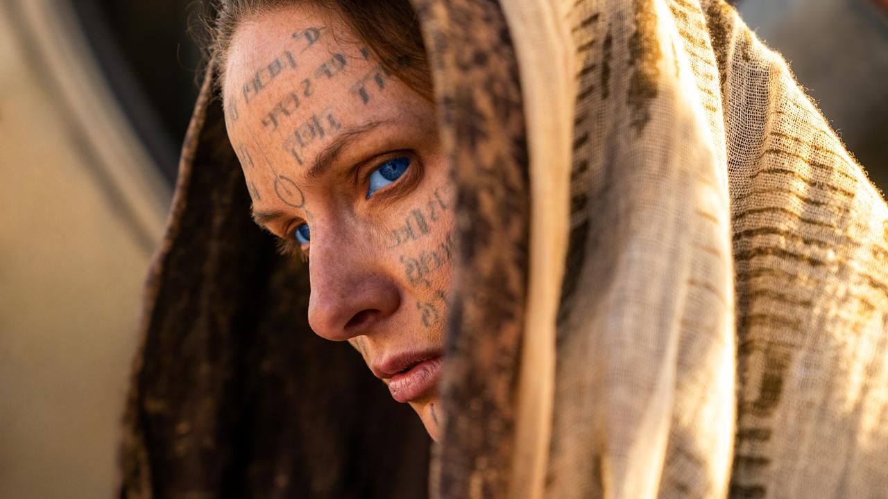 Rebecca Ferguson als Lady Jessica Atreides mit einem Umhang über ihren Kopf blickt sie mit ihren blauen Augen ernst, in ihrem orangefarben geschminkten Gesicht sind Worte in fremder Schrift geschrieeben
