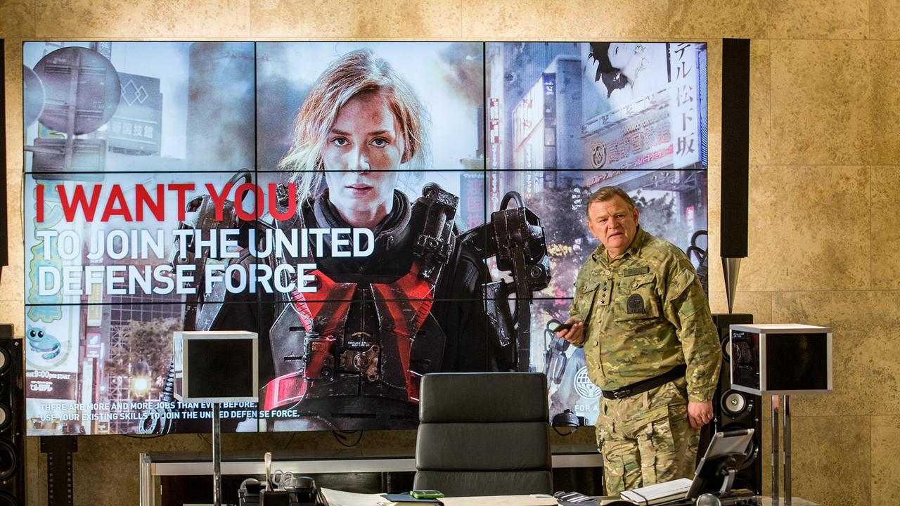 © ORF/Warner Bros. Pictures Brendan Gleeson steht als General Brigham in seiner Uniform vor einem großem LED-Schirm, auf dem Emily Blunt als Rita zu sehen ist - mit der Aufschrift I WANT YOU TO JOIN THE UNITED DEFENSE FORCE