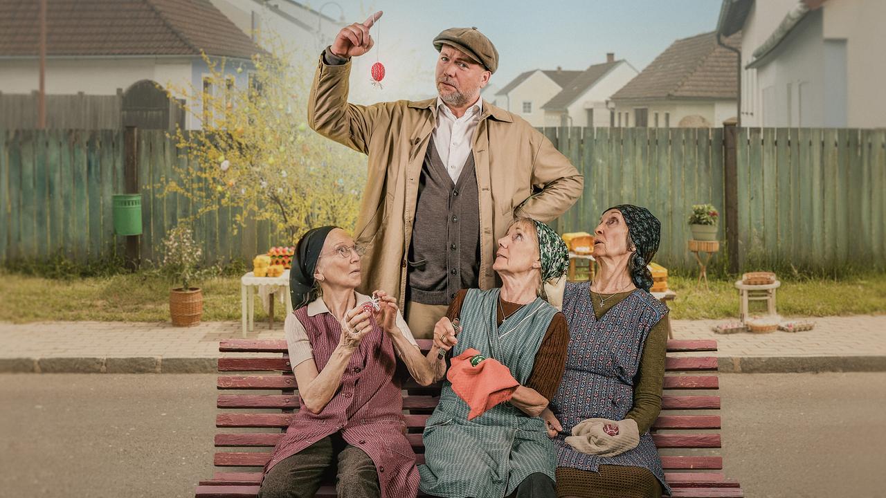 Sifkovits steht hinter den drei älteren Frauen Resl Grandits, Baba Sifkovits und Hilda Resetarits auf einer Parkbank in einer burgenländischen Dorflandschaft und hält einen kleines rotes bemaltes Osterei in die Höhe.