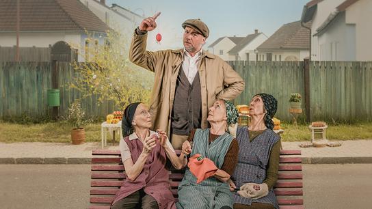 Sifkovits steht hinter den drei älteren Frauen Resl Grandits, Baba Sifkovits und Hilda Resetarits auf einer Parkbank in einer burgenländischen Dorflandschaft und hält einen kleines rotes bemaltes Osterei in die Höhe.