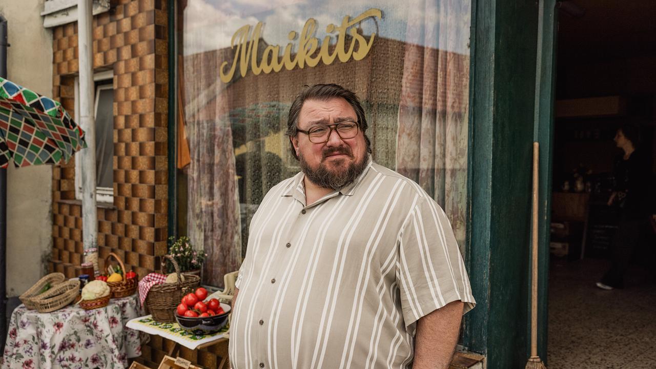 Der korpulente Franz Maikits steht vor seiner kleinen Greißler mit Obstkisten und Gemüseregalen.