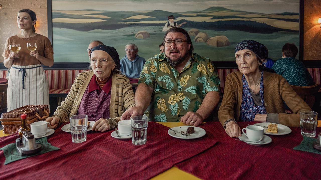 Resl Grandits, Franz Maikits und Hilda Resetarits sitzen in einem Wirtshaus an einem Tisch mit Kaffee und Mehlspeisen und schauen überrascht nach vorne, während eine Kellnerin Getränke serviert.