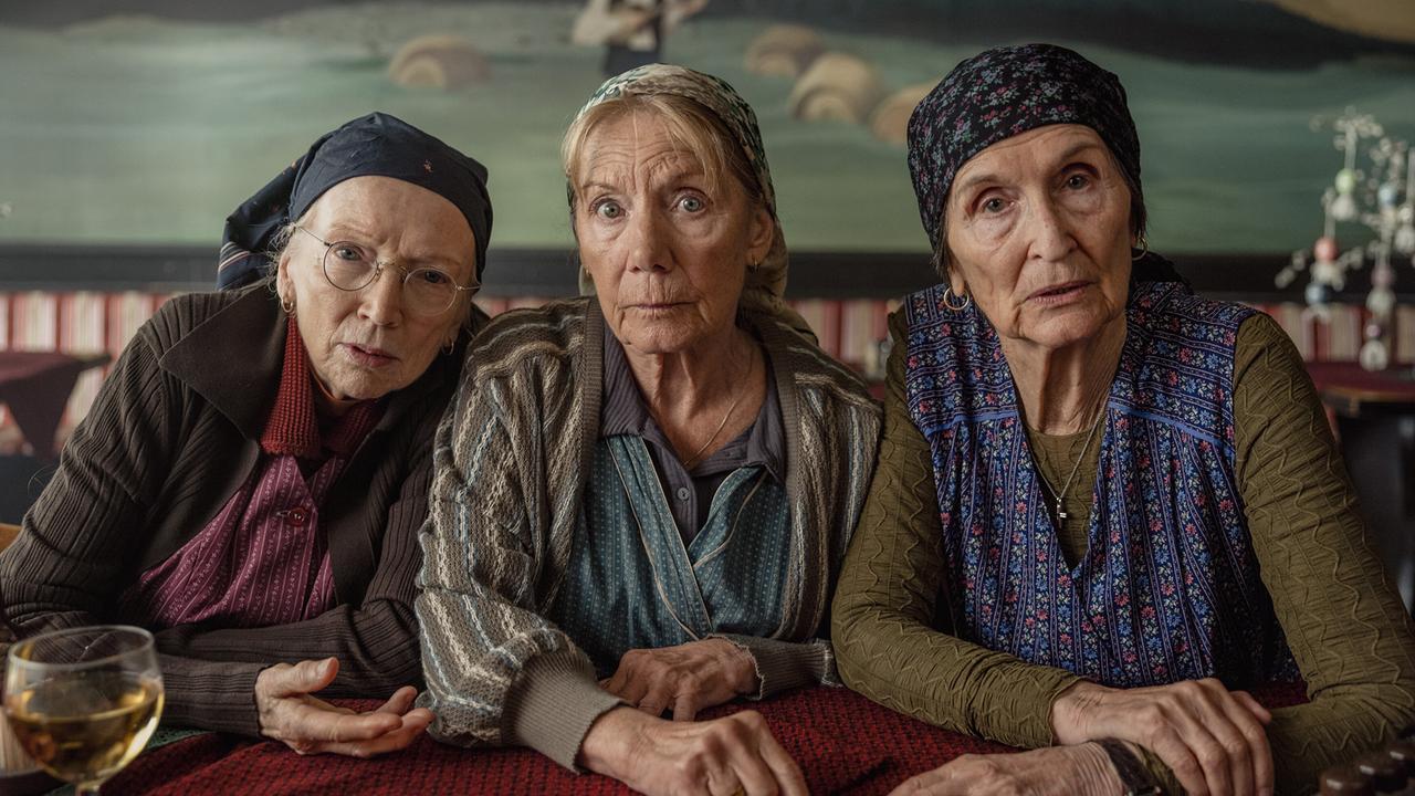 Die drei ältere Frauen Resl Grandits, Baba Sifkovits und Hilda Resetarits sitzen dicht nebeneinander an einem Wirtshaustisch und schauen aufmerksam nach vorne.