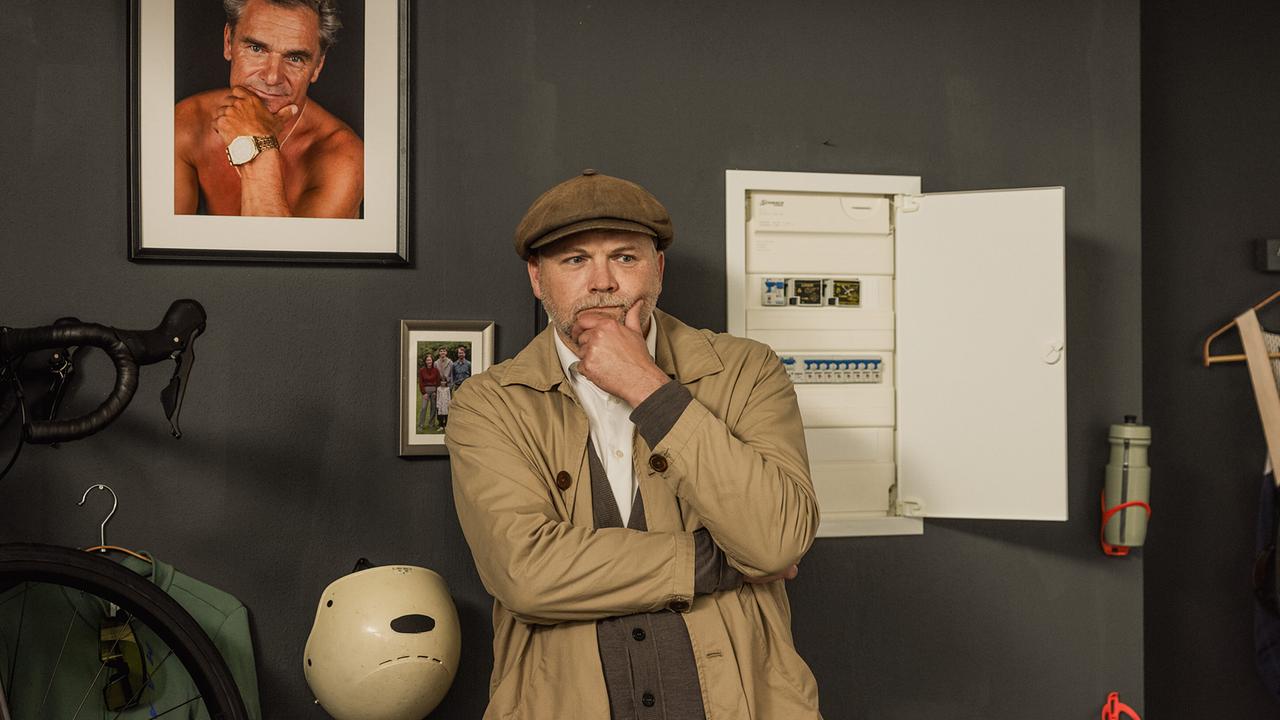 Sifkovitsn steht nachdenklich in einem Raum an einer Wand neben einem Stromkasten, Radfahrutensilien und gerahmten Fotos an der Wand.