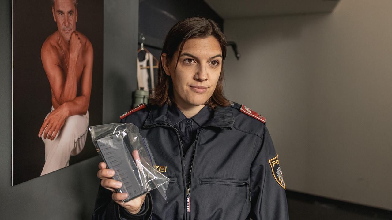 Polizistin Kampitsch hält in einem Innenraum ein sichergestelltes Smartphone in einem durchsichtigen Beutel hoch.