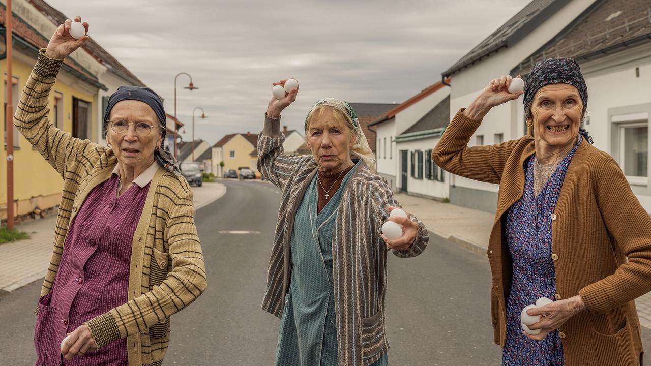 Resl Grandits, Baba Sifkovits und Hilda Resetarits stehen auf einer Dorfstraße und scheinen weiße Eier mit erhobenen Armen werfen zu wollen.