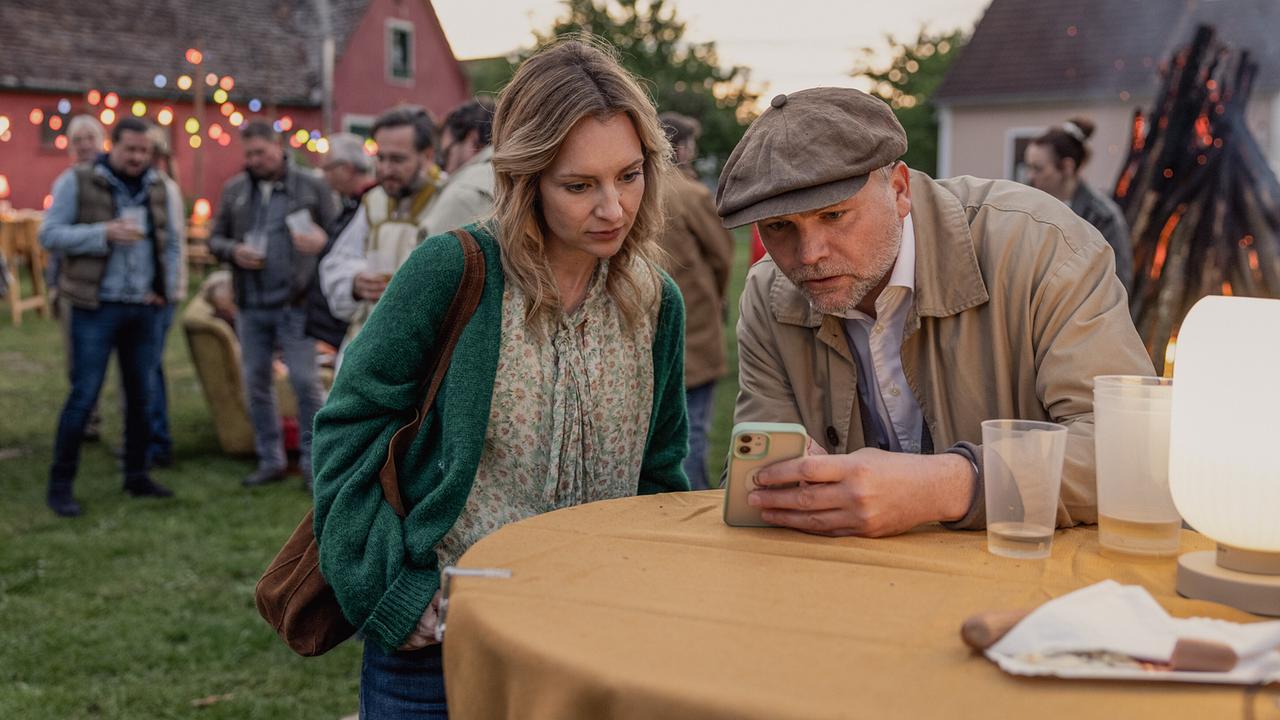 Bei einem Gartenfest beugen sich Lisa Sifkovits und Sifkovits über ein Smartphone auf einem Stehtisch, und schauen sich darauf etwas an.