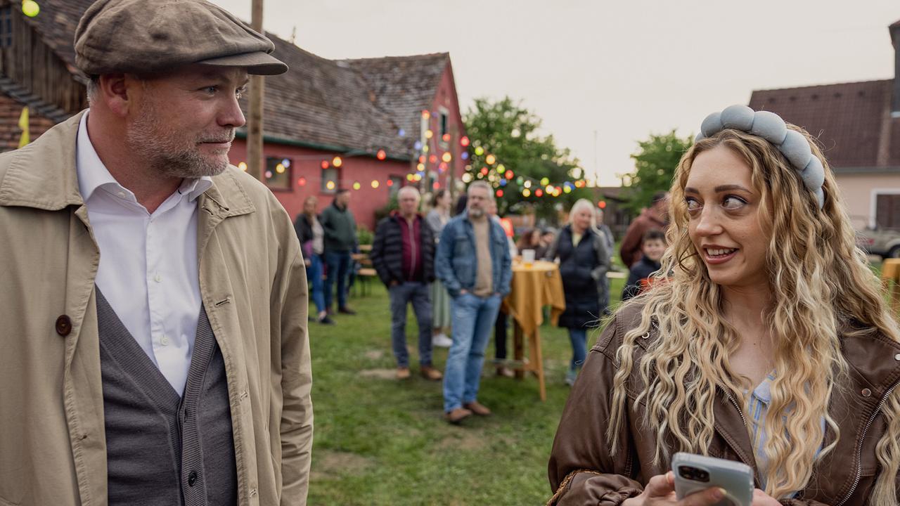 Sifkovits und die junge Maja Janisch stehen untertags im Freien bei einer Feier, während sie auf ihr Handy schaut und Menschen im Hintergrund an Stehtischen plaudern.