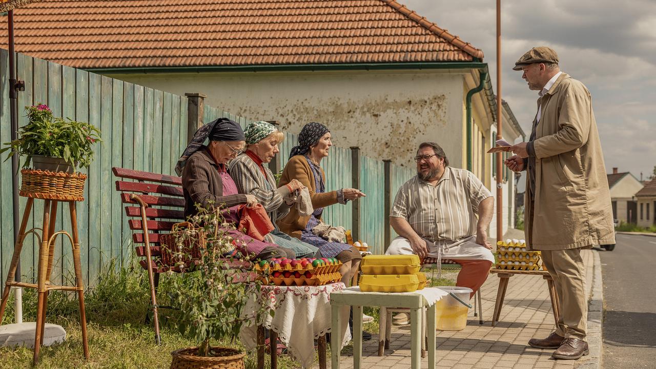 Resl Grandits, Baba Sifkovits, Hilda Resetarits und Franz Maikits sitzen auf dem Gehsteig vor einem Haus an einem improvisierten Verkaufsstand mit Eierkartons, während Sifkovits vor ihnen steht und etwas notiert.