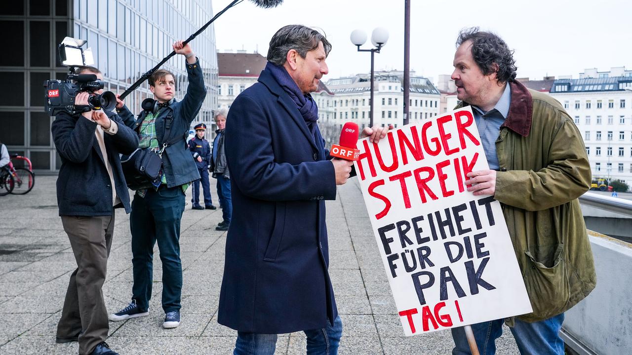 "Eigentlich sollten wir": Stefan Steindl (Thomas Mraz) steht vor der Kripo mit einer Protesttafel, auf der „Hungerstreik! Freiheit für die PAK“ steht . ORF- Reporter Serge (Florian Pissecker) interviewt ihn.