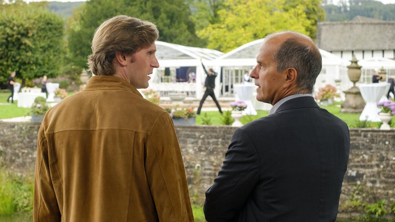 Lasse und Event-Manager Dieter Salzmann stehen im Schlossgarten nebeneinander und schauen einander an, im Hintergrund ist ein Tänzer vor Festzelten zu sehen