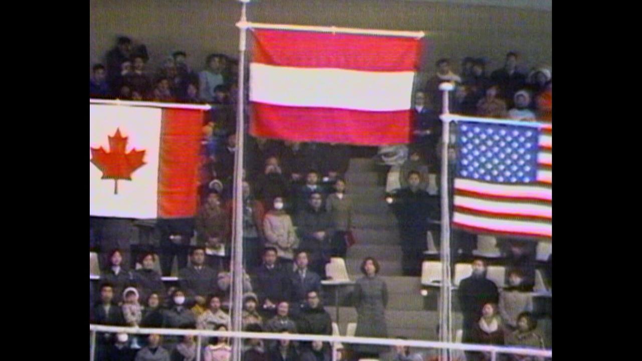 © ORF/ORF Sport Eine große Zuschauermenge sieht zu, wie die kanadische, österreichische und US-amerikanische Flagge bei einer Siegerehrung gehisst werden.