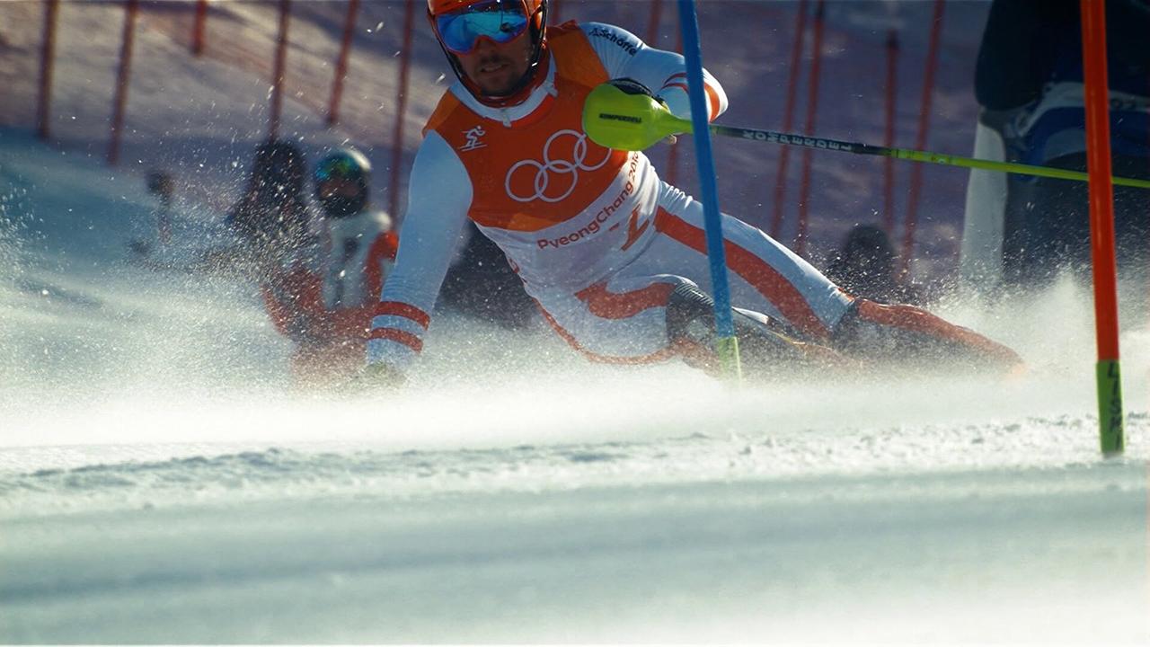 © ORF/ORF Sport Marcel Hirscher im Rennanzug fährt bei einem olympischen Slalomrennen eng an einem Tor vorbei und wirbelt dabei Schnee auf.