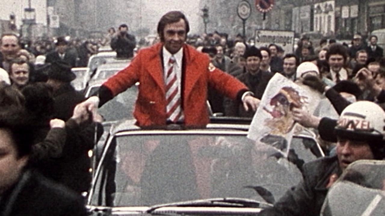 © ORF/ORF Sport Karl Schranz in roter Jacke fährt stehend im offenen Auto durch eine jubelnde Menschenmenge, die ihn am Straßenrand begeistert empfängt.