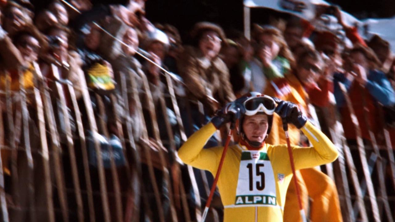 © ORF/ORF Sport Franz Klammer im gelben Skianzug jubelt vor einer großen Zuschauermenge hinter Absperrungen.