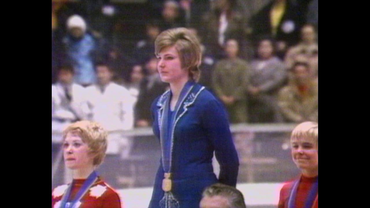 © ORF/ORF Sport Trixi Schuba steht auf einem Siegerpodest und trägt eine Goldmedaille, während das Publikum im Hintergrund zusieht.