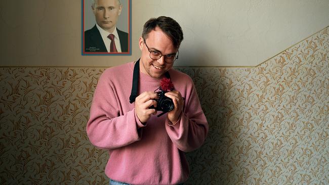 Pawel Talankin mit einer Kamera in den Händen, lächelt freundlich aus dem Bild, hinter ihm an der Wand ein Porträt des russichen Präsidenten Wladimir Putin