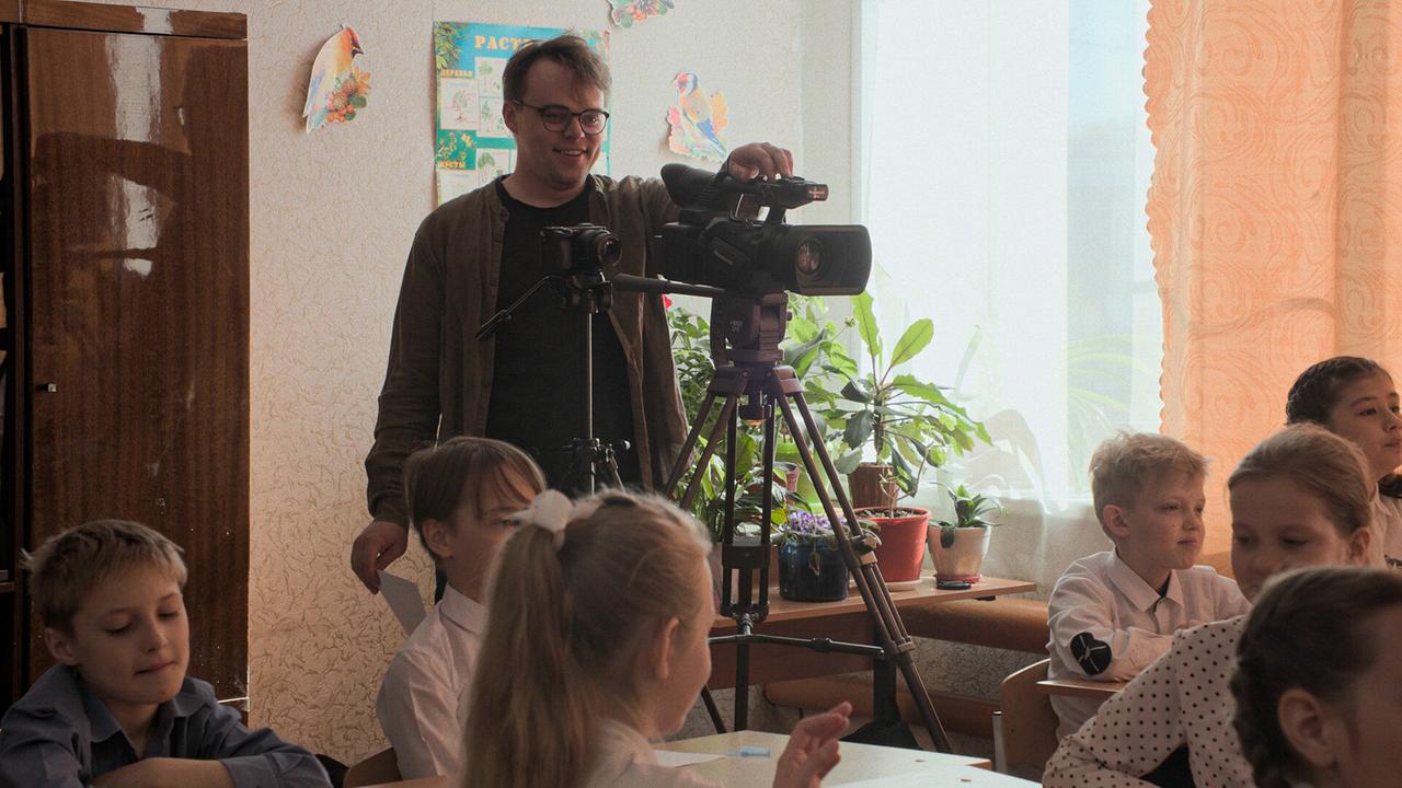 Pawel Talankin im Klassenzimmer, hinter der auf einem Stativ stehenden Videokamera, vor ihm sitzen die Schulkinder in den Reihen, dahinter ein Tisch mit ZImmerpflanzen