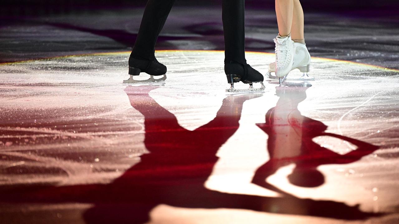 Foto der Füße und Eislaufschuhe eines Eiskunstlaufpaars  auf einer beleuchteten Eisfläche, deren farbiges Licht ihre Schatten auf dem Eis erscheinen lässt.
