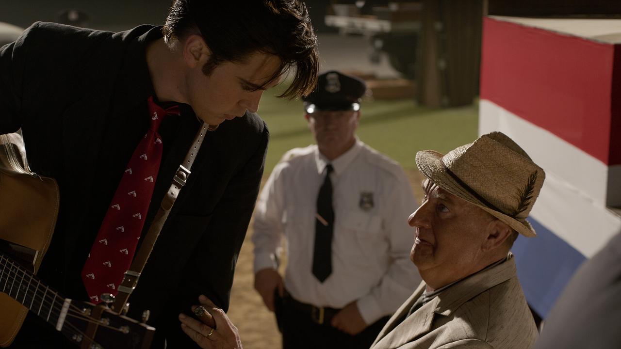 Ein schwarz gekleideter, junger Mann mit Gitarre wendet sich zu einem älteren Mann mit Zigarre in der Hand, im Hintergrund steht ein Streifenpolizist
