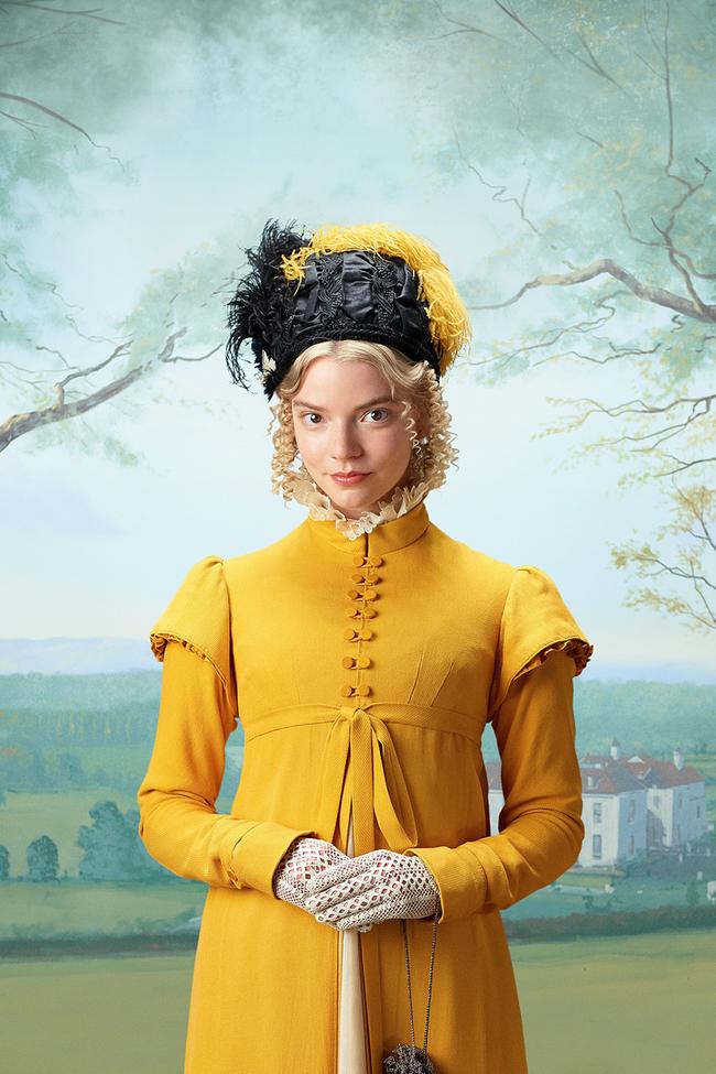 Anya Taylor-Joy steht als Emma Woodhouse in einem langen, gelben Kleid und passendem Hut mit schwarzer Verzierung vor einer gemalten Landschaft mit Feldern und einem Herrenhaus