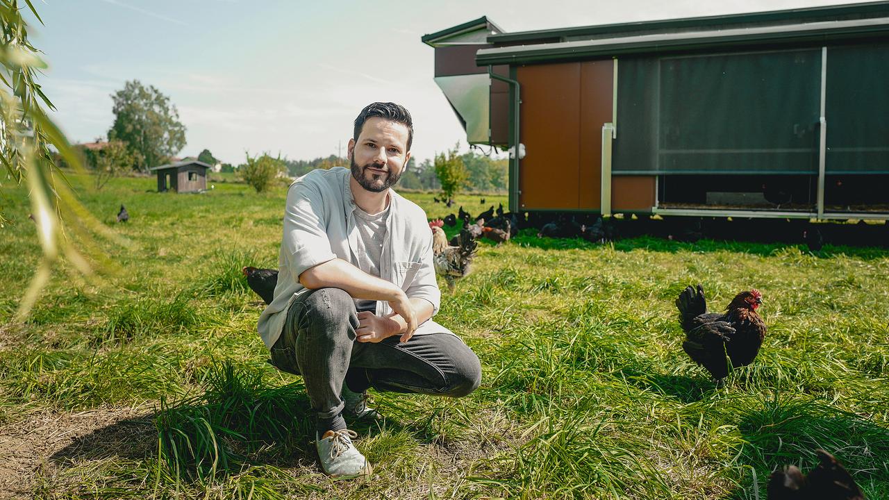 © ORF/WDR/Sebastian Wallner Niko Rittenau, ein junger, bärtiger Mann in Jeans, T-Shirt und offenem Hemd hockt auf einer Wiese vor einem mobilen Hühnerstall, umgeben von frei laufenden Hühnern.