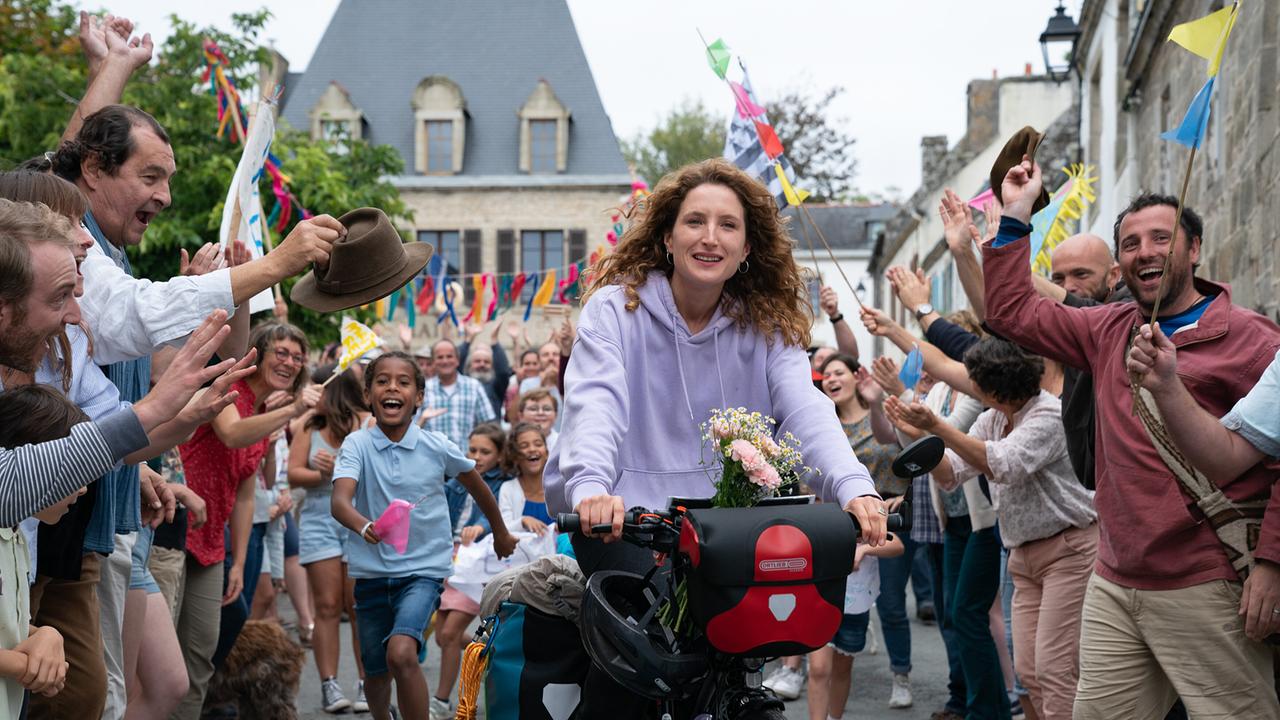 © ORF/Silkwayfilms/Stephanie Branchu Eine junge Frau fährt bei einem Dorffest mit einem mit Blumen geschmückten Rad, jubelnde Menschen an der Straßenseite, Kinder laufen lachend hinter ihr her