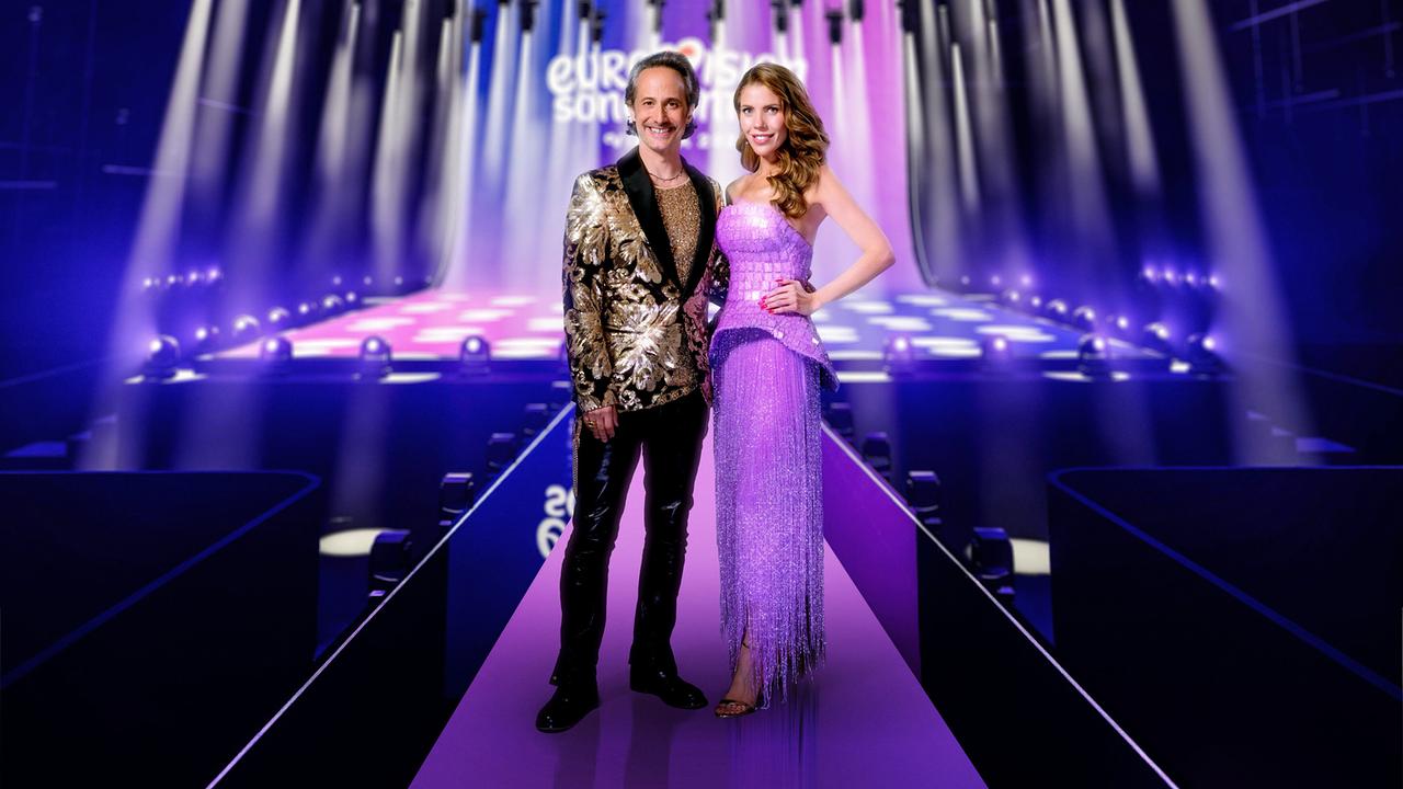 Michael Ostrowski und Victoria Swarovski stehen in eleganten glitzernden Outfits auf einer bunt beleuchteten Bühne mit "Eurovision Song Contest"-Schriftzug im Hintergrund, umgeben von vielen Scheinwerfern und einem modernen Bühnenset.