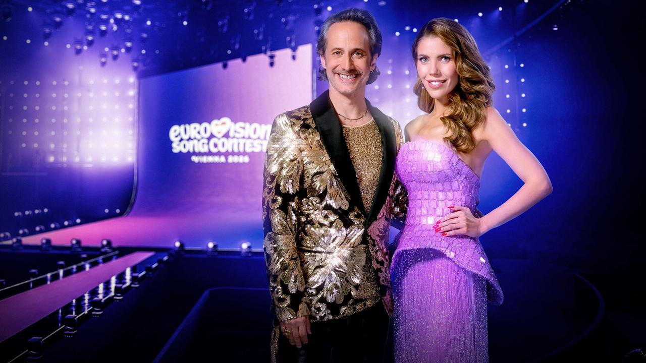 Michael Ostrowski und Victoria Swarovski stehen in eleganten glitzernden Outfits auf einer bunt beleuchteten Bühne mit "Eurovision Song Contest"-Schriftzug im Hintergrund, umgeben von vielen Scheinwerfern und einem modernen Bühnenset.