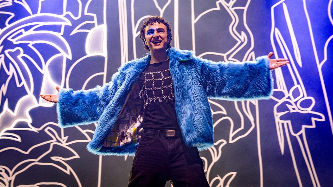 COSMÓ in einer lauen Kunstfelljacke mit ausgebreiteten Armen auf der Bühne vor einem grafisch beleuchteten Hintergrund posiert.