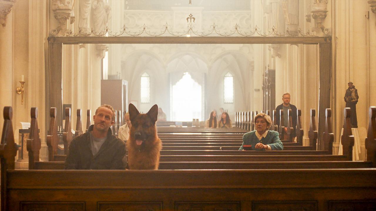 Maximilian Brückner und Rex sitzen in einer Kirchenbank in einem hellen Kirchenraum, der mit vier weiteren Besucher gefüllt ist.
